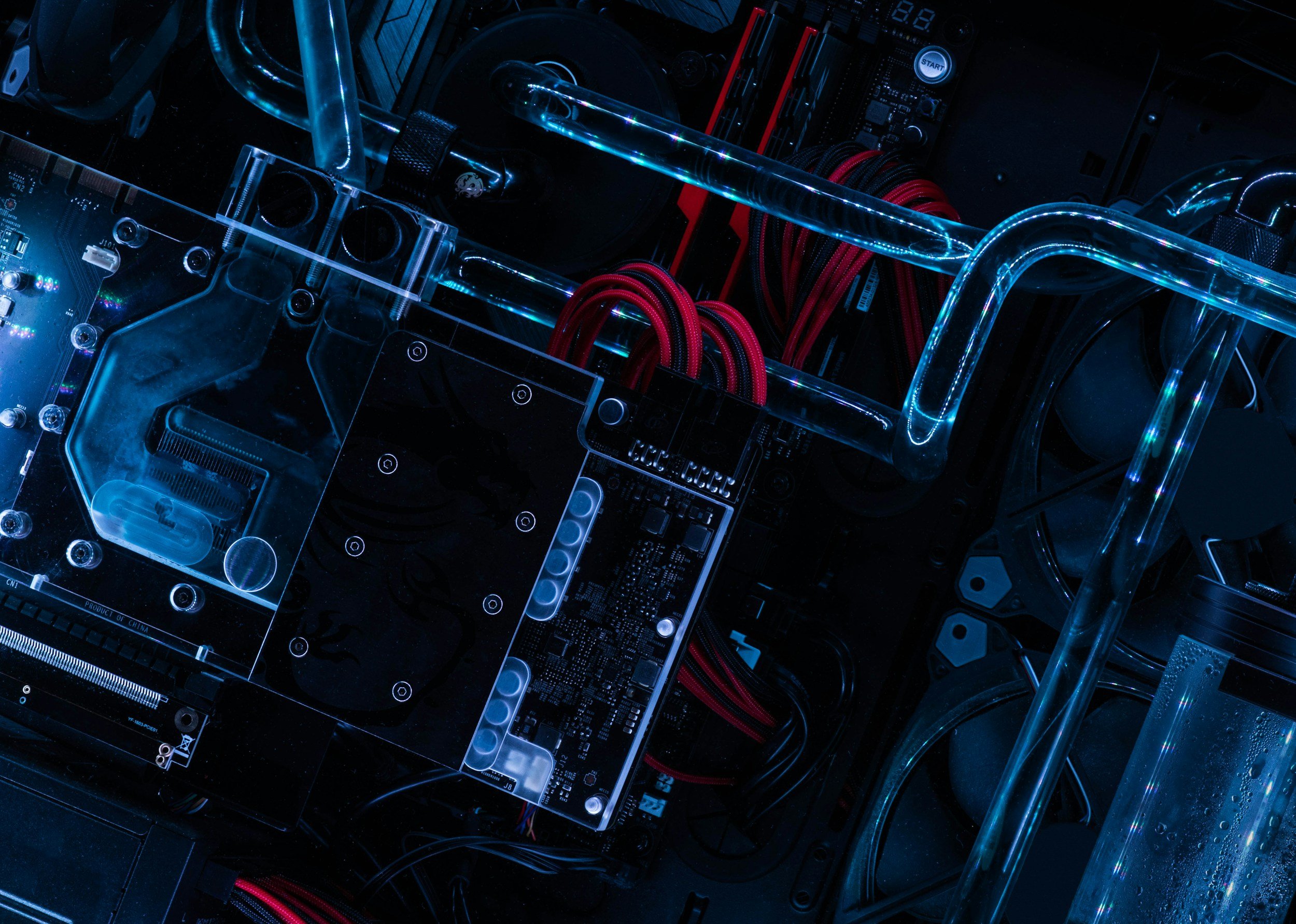Close-up view of a custom-built computer with transparent water cooling tubes, circuit boards, and red and black cables illuminated with blue lighting.